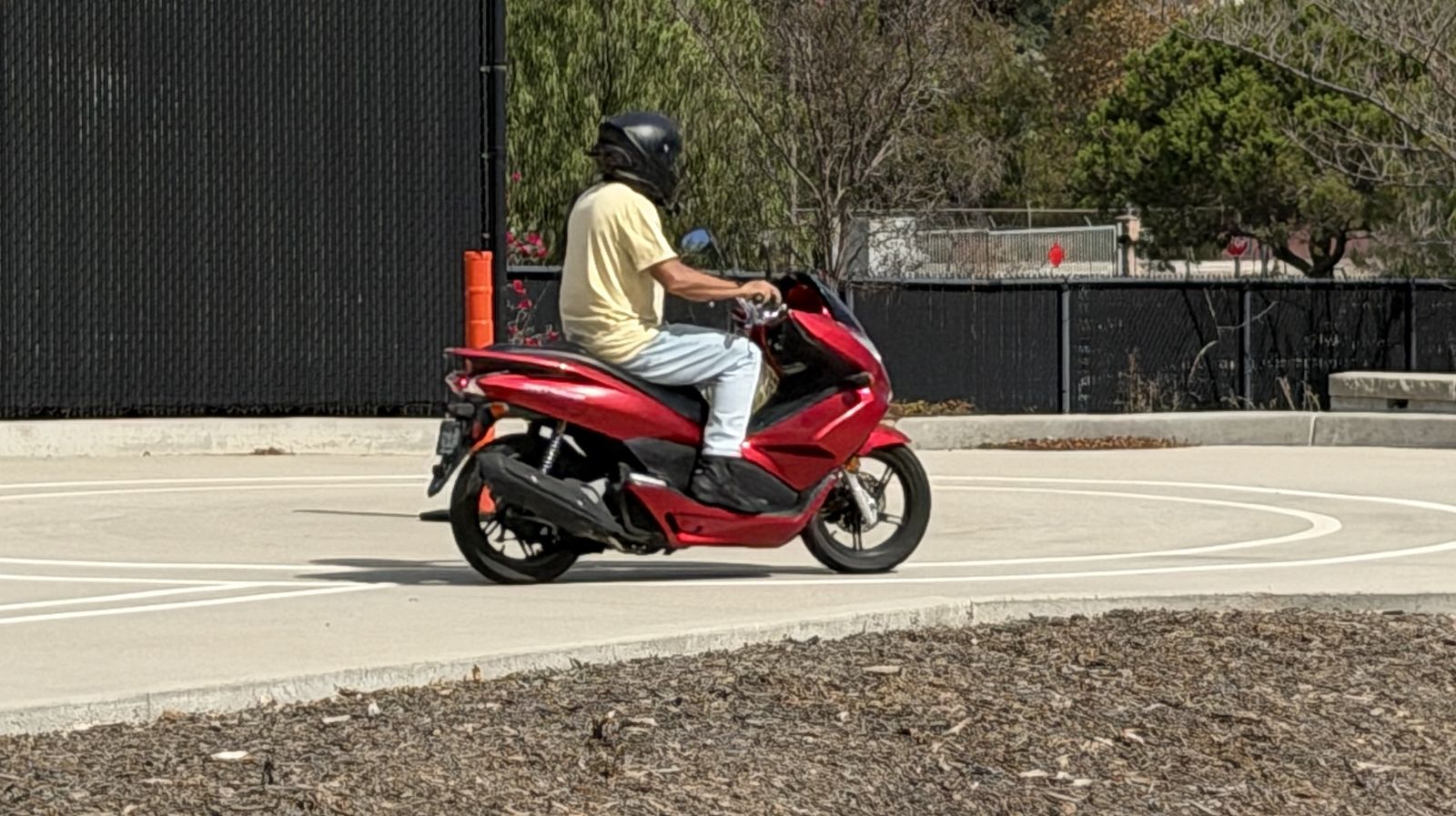 Rider in a black helmet on a red Honda PCX scooter, riding the painted circles of a motorcycle skills test course