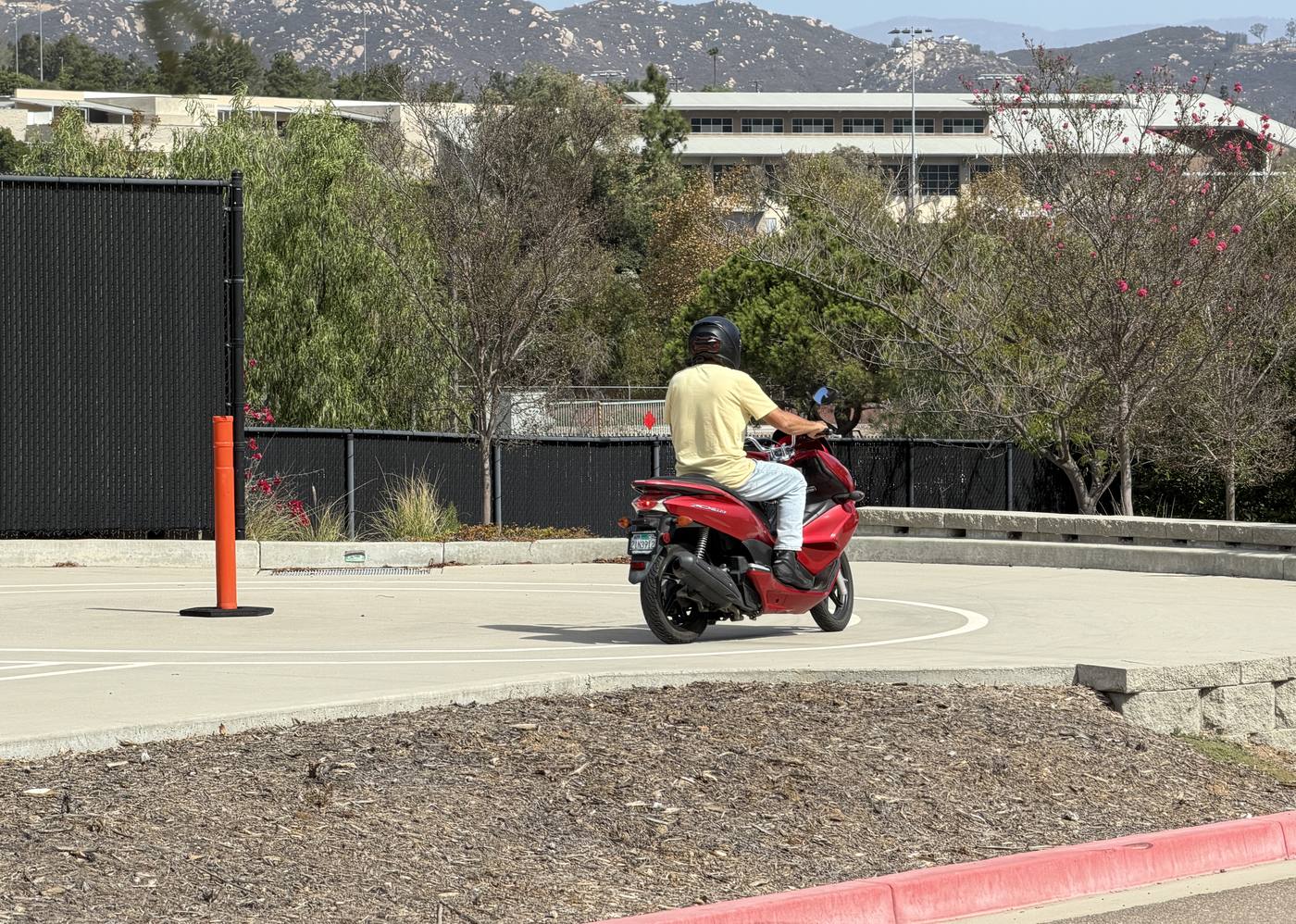 Rider on a red Honda PCX scooter leaning through a curve on a motorcycle skills test course, past an orange marker post